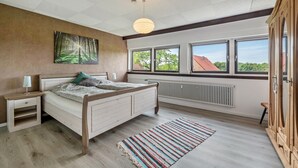 Une chambre à coucher comprenant un lit, une table de chevet, une commode, un radiateur, des fenêtres et une vue sur l’extérieur.