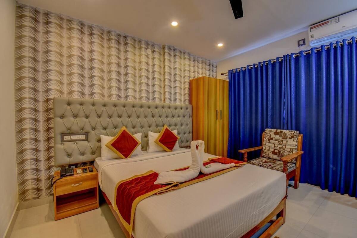 A hotel room with a tufted headboard, a bed with white bedding and red accents, a wooden nightstand, a chair, and a large window with blue curtains.