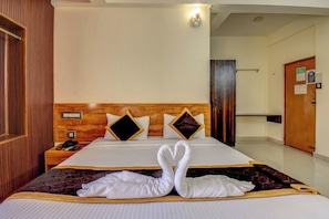 Habitación de hotel con cabecera de madera, cama con sábanas blancas, dos almohadas, mesita de noche con teléfono y lámpara de pared.