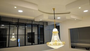 A chandelier with a gold chain and crystal design, hanging in a room with large windows and recessed ceiling lights. - ATITHI COMFORTS (Somvarpet)