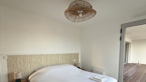 A bedroom with a large bed, a nightstand, a lamp, and a view of a wooden deck through a glass door. - Apartment with terrace – Close to the sea and Royan city center (Royan)