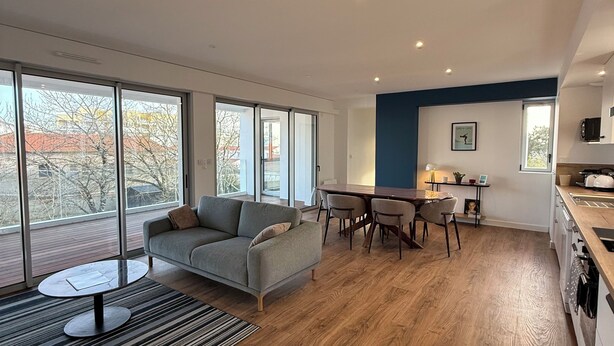 A modern living space with a sofa, dining table, and kitchen area, featuring large windows and wooden flooring. - Apartment with terrace – Close to the sea and Royan city center (Royan)