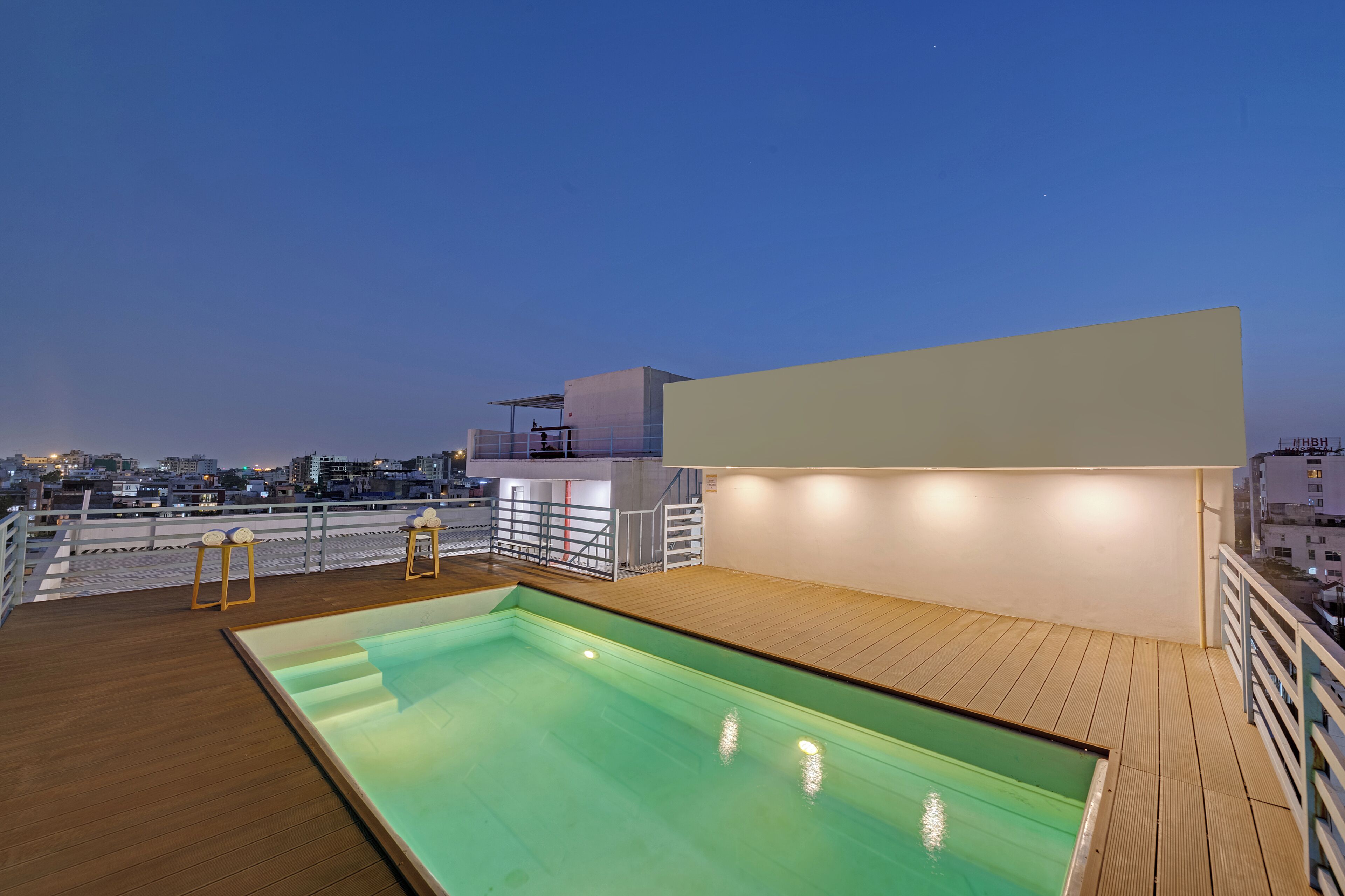 Une piscine sur le toit, avec une terrasse en bois, un mur blanc et une vue sur le paysage urbain au crépuscule.