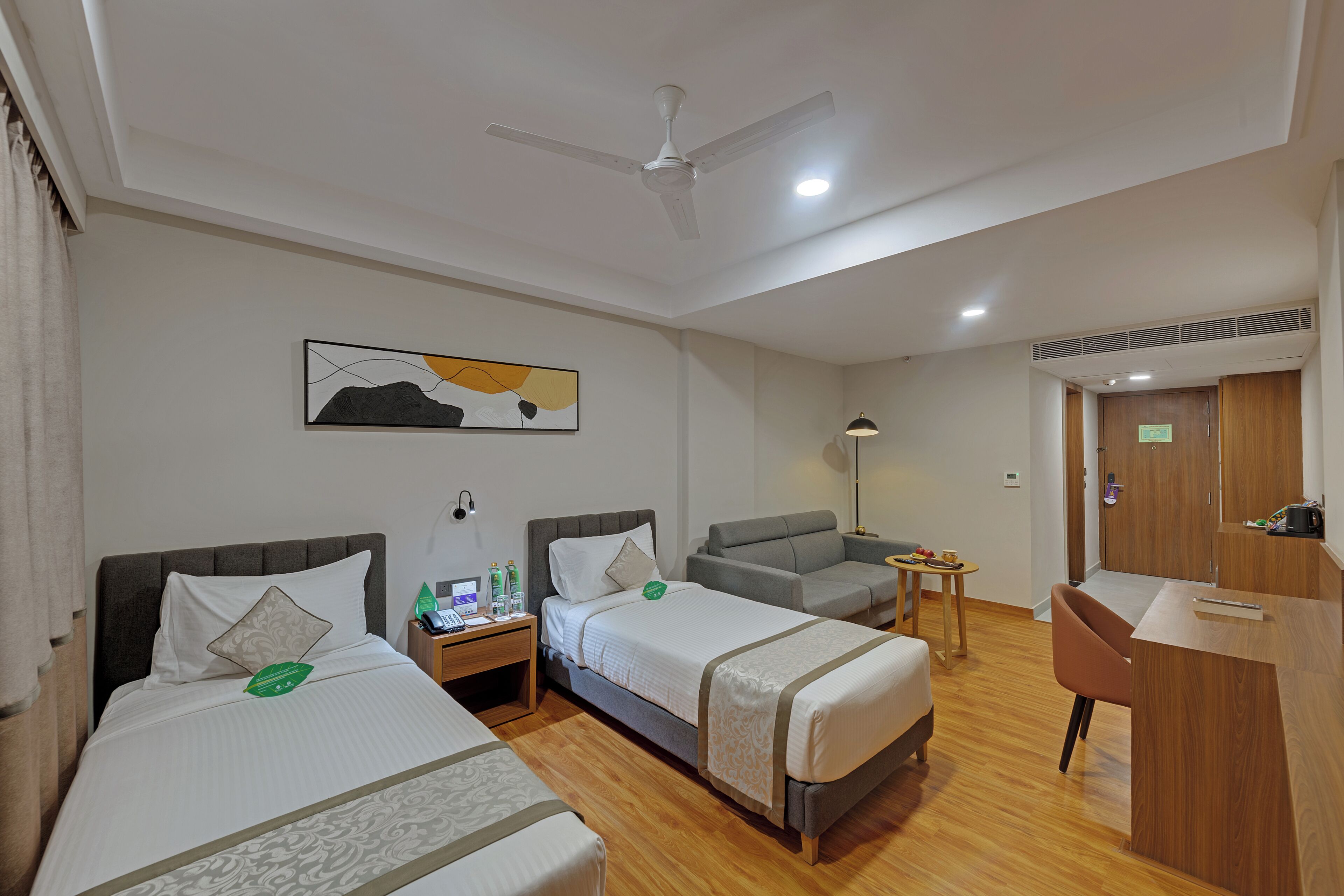 Une chambre d’hôtel avec deux lits, un bureau, une chaise, un ventilateur de plafond, une table de chevet, une lampe et un tableau au mur.