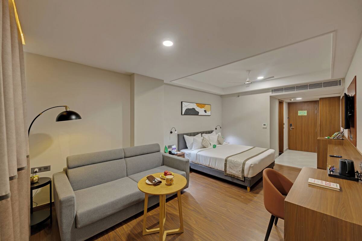 A hotel room with a bed, a sofa, a desk, a chair, a table, a lamp, and a painting on the wall.