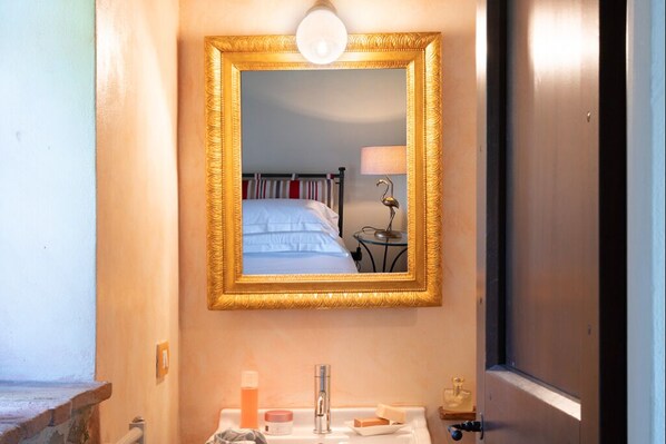 A bathroom with a mirror, sink, and a bed visible in the reflection.