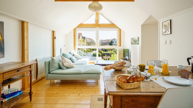 A cozy room with a wooden ceiling, a sofa, a dining table with breakfast items, and a view through a large window. - Guest Room 'Auguste' with Shared Pool (Plobsheim)