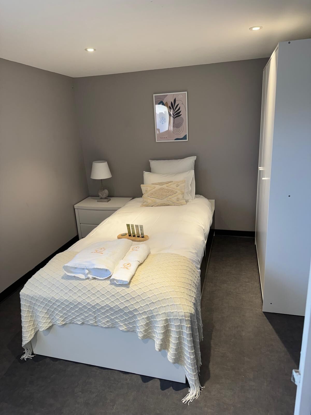 Cama bem arrumada com colcha, luminária de cabeceira e mesa pequena com artigos de higiene pessoal.
