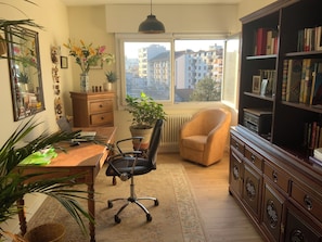 Ein Zimmer mit Schreibtisch, Stuhl, Bücherregal, Topfpflanzen und einem Fenster mit Stadtblick.