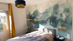 Ett sovrum med en säng, ett nattduksbord, en lampa, en vägg med abstrakt konst, ett fönster med gardiner och utsikt över byggnader utanför.