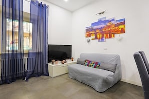 A living room with a grey sofa, a flat-screen TV, a white side table, and a window with blue curtains. - Loving Sweet Roma by Rentbeat (Roma)
