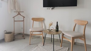 Een kamer met twee houten stoelen, een klein rond tafeltje met een fles en glazen, een aan de muur bevestigde televisie en een kledingrek met hangers.