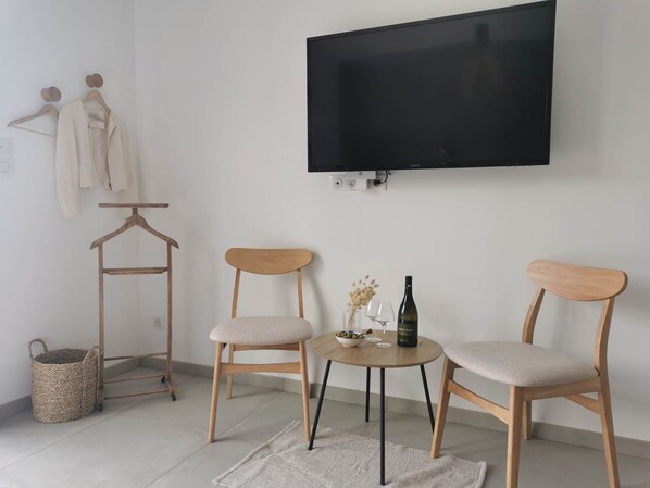 A room with two wooden chairs, a small round table with a bottle and glasses, a TV mounted on the wall, and a clothing rack with hangers.