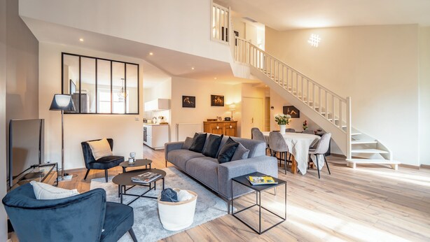 Een moderne woonkamer met een grijze bank, zwarte fauteuils, een salontafel en een eethoek met tafel en stoelen. Een trap met een witte leuning leidt naar een hoger gelegen verdieping.