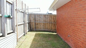 Un patio trasero con valla de madera, pared de ladrillo y una ventana con un objeto verde en el alféizar.