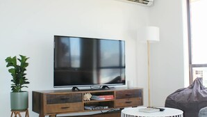 Una sala de estar con televisor de pantalla plana, mueble de madera para equipos electrónicos, planta en maceta, lámpara de pie, mesa de centro redonda blanca y un sillón orejero.