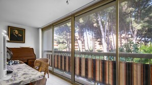 A room with a dining table, chairs, a piano, a radiator, and a large window with a view of trees and buildings. - Beautiful 2-Bedroom Apartment in Green Estate with Shared Pool, Balcony & Wi-Fi (Six-Fours-les-Plages)