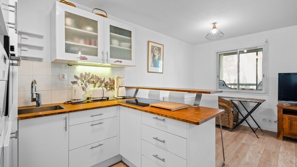 A modern kitchen with white cabinets, a wooden countertop, and a dining area with a TV and window. - Beautiful 2-Bedroom Apartment in Green Estate with Shared Pool, Balcony & Wi-Fi (Six-Fours-les-Plages)