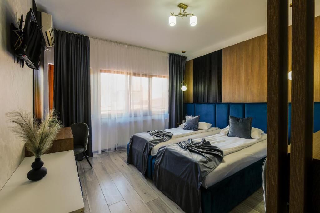 Une chambre d’hôtel avec deux lits, un bureau, une chaise, une plante en pot et une grande fenêtre avec des rideaux.