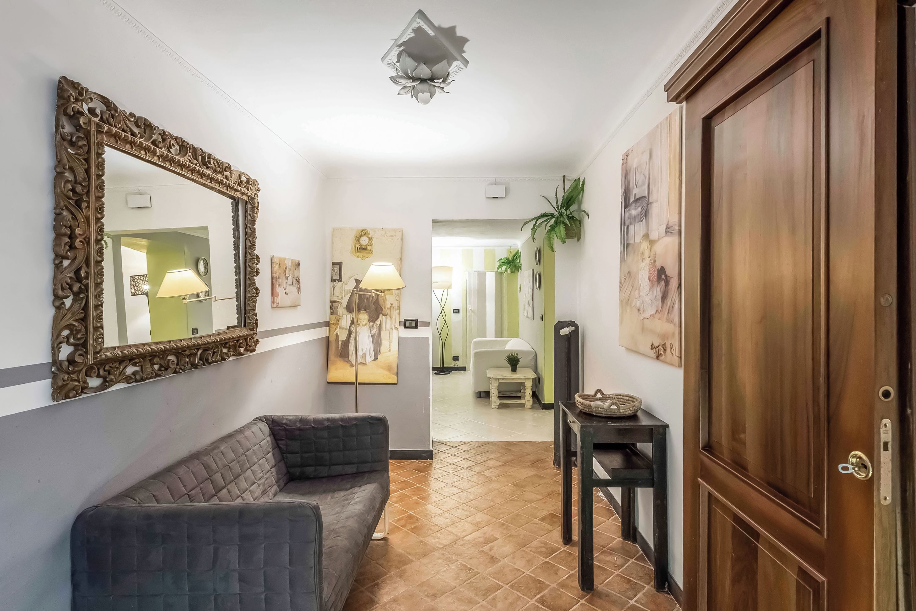 A hallway with a dark-colored sofa, a large ornate mirror, a wooden door, and a tiled floor.