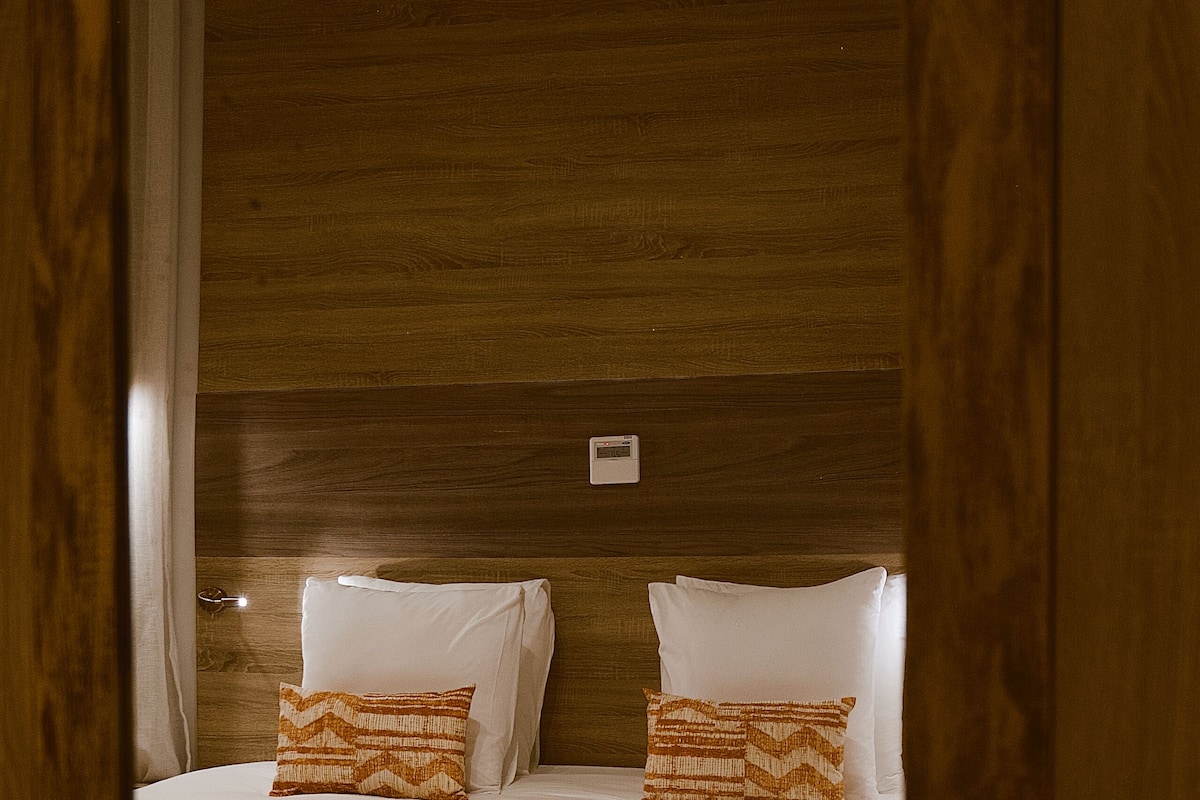 Habitación de hotel con cabecera de madera, cama con sábanas blancas y almohadas estampadas, un banco al pie de la cama y una mesita de noche con lámpara.