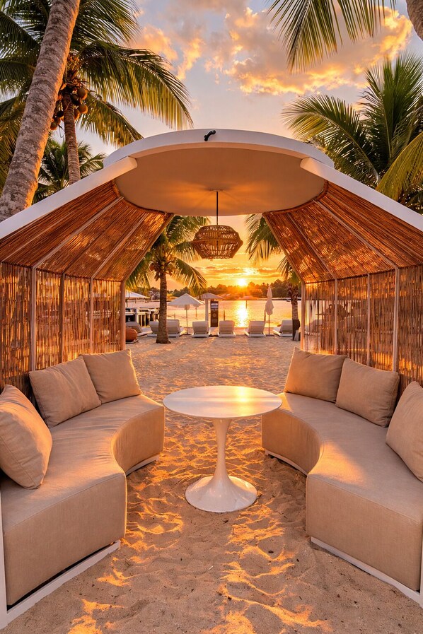 A beach cabana with a round table and two sofas, surrounded by palm trees and a sunset view.
