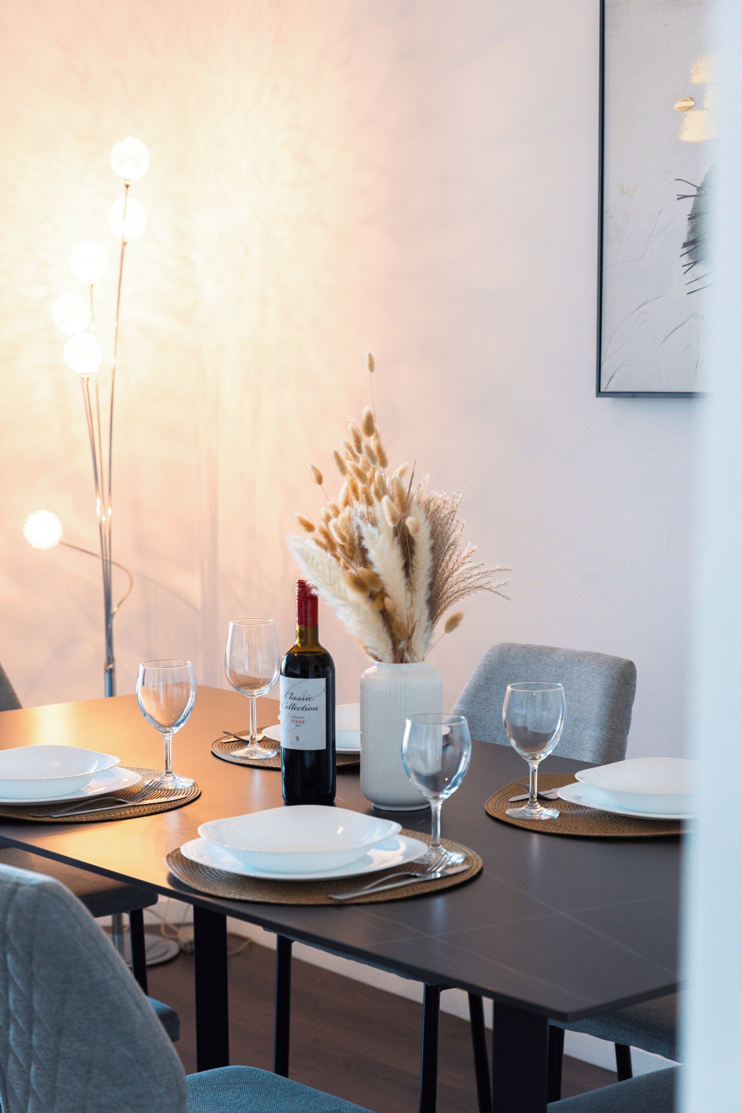Uma mesa de jantar posta com pratos, copos e uma garrafa de vinho, com um vaso de capim-dos-pampas e uma luminária moderna ao fundo.
