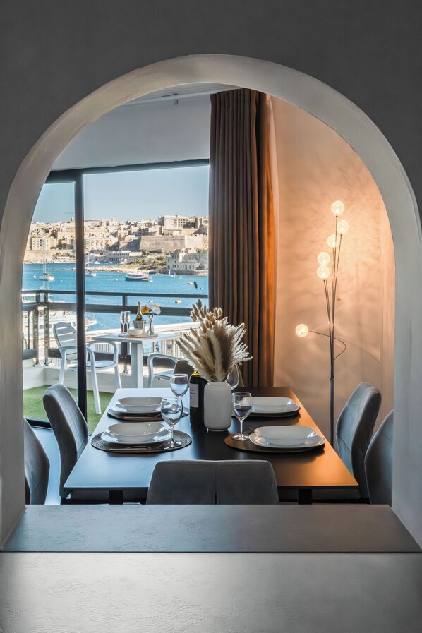 A dining area with a table set for two, a vase with flowers, and a view of the sea and buildings. - Sliema Zara (Sliema)
