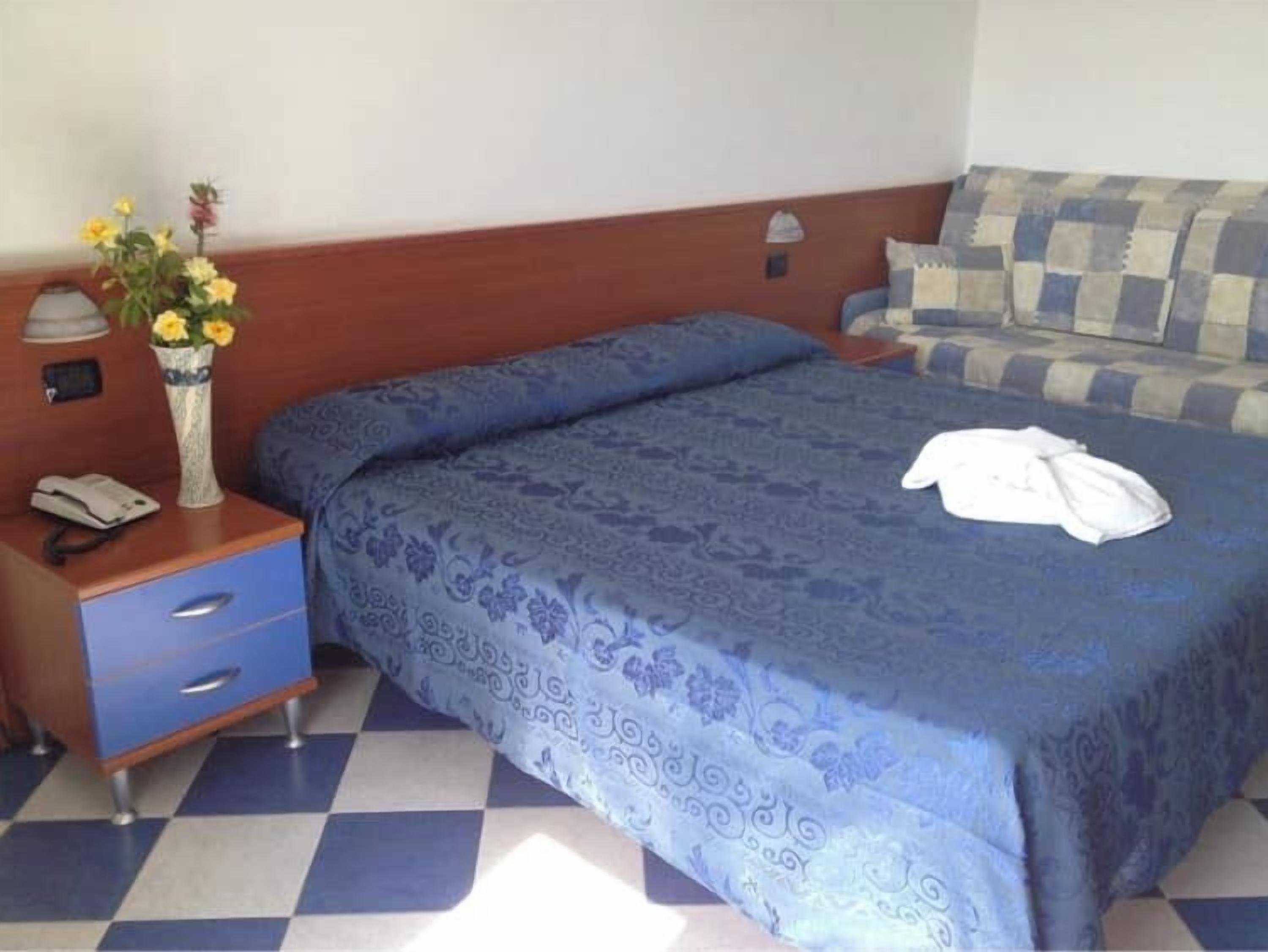 A hotel room with a checkered floor, a bed with a blue patterned bedspread, a bedside table with a phone, and a vase with flowers.