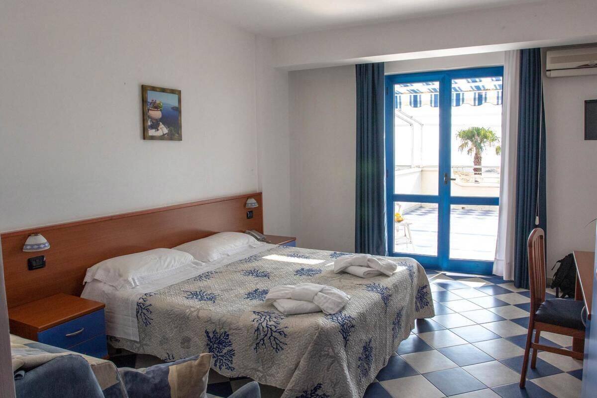 Ein Hotelzimmer mit Bett, Schreibtisch, Stuhl, Nachttisch, einer Wandlampe, einem Fenster mit Vorhängen und einer zur Außenwelt führenden Tür.