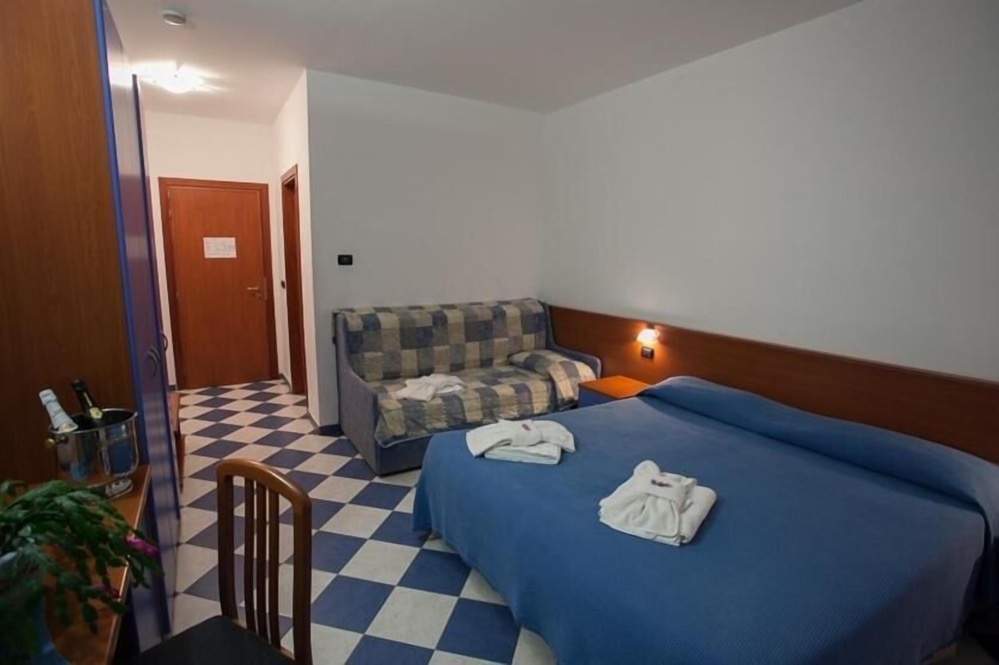 A hotel room with a checkered floor, a bed with blue bedding, a sofa with a checkered cushion, a small table with a bottle and glasses, and a door with a sign.
