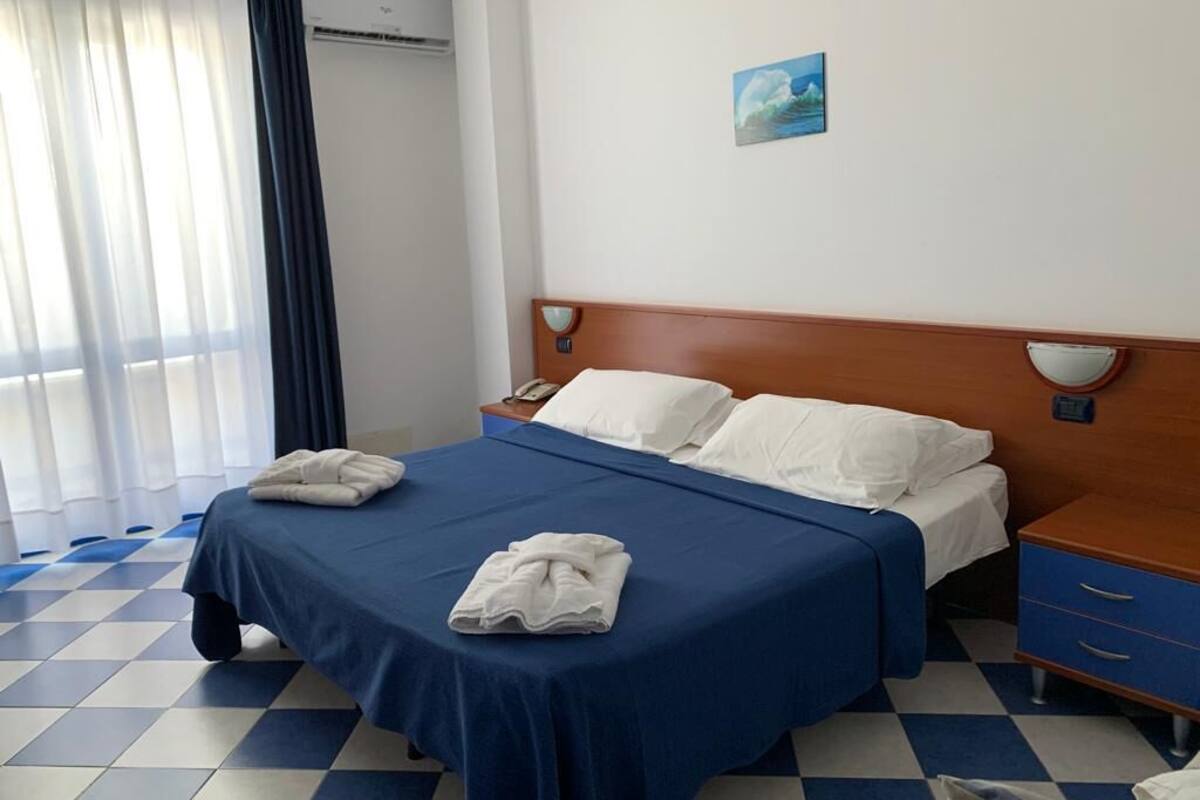 Ein Hotelzimmer mit Schachbrettmuster-Boden, einem Bett mit blauer Bettdecke, weißen Kissen, einem Holzkopfteil, einem Nachttisch mit Telefon und einem Gemälde an der Wand.
