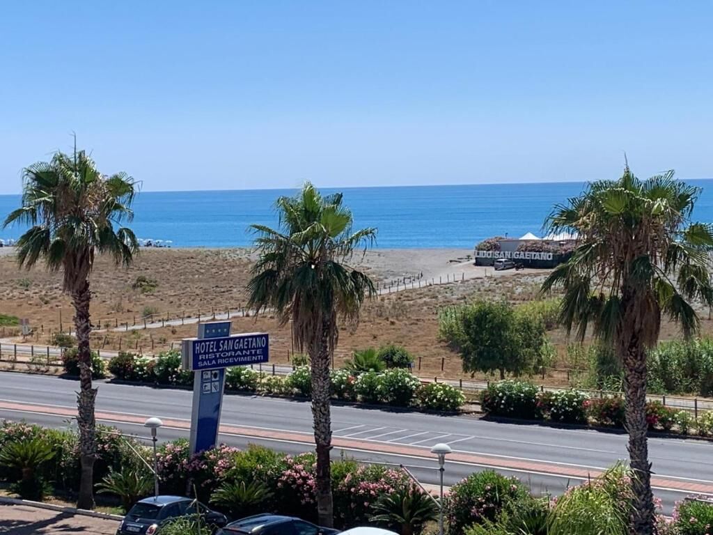 Eine Küstenstraße mit einem Schild für Hotel San Gaetano, Palmen und einem Strand im Hintergrund.