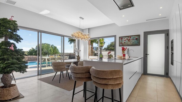A modern kitchen with a large island, bar stools, a potted tree, and a view of the outdoors. - Villa PinkBirdie de Luxe. Frontline Golf Las Colinas Piscine Rooftop Bali Lounge (Dehesa de Campoamor)