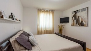 Ein Schlafzimmer mit Bett, Fernseher, einem Gemälde und einem Fenster mit Vorhängen.