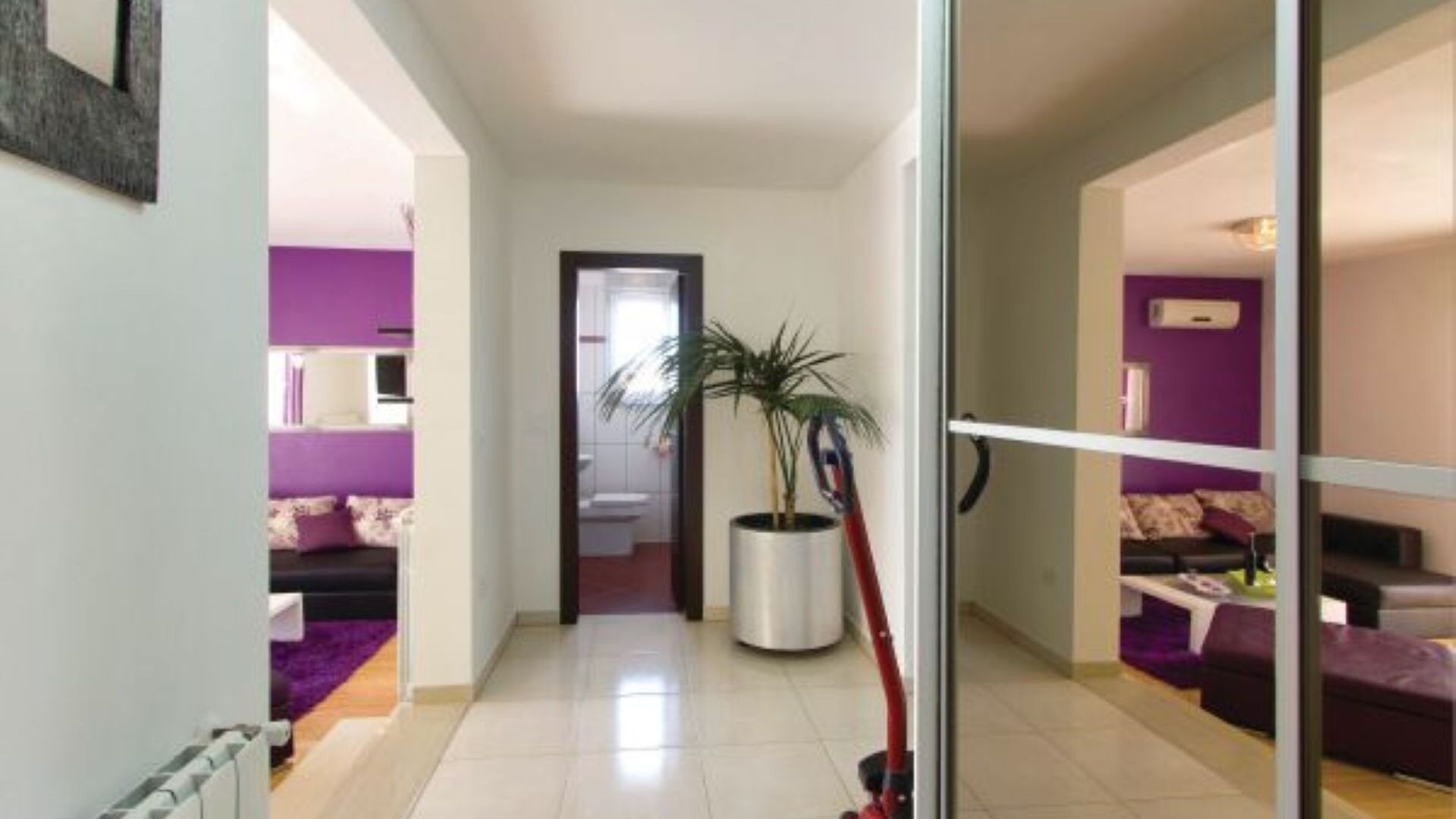 A hallway with a potted plant, a door leading to a bathroom, and a living area with purple walls and furniture.