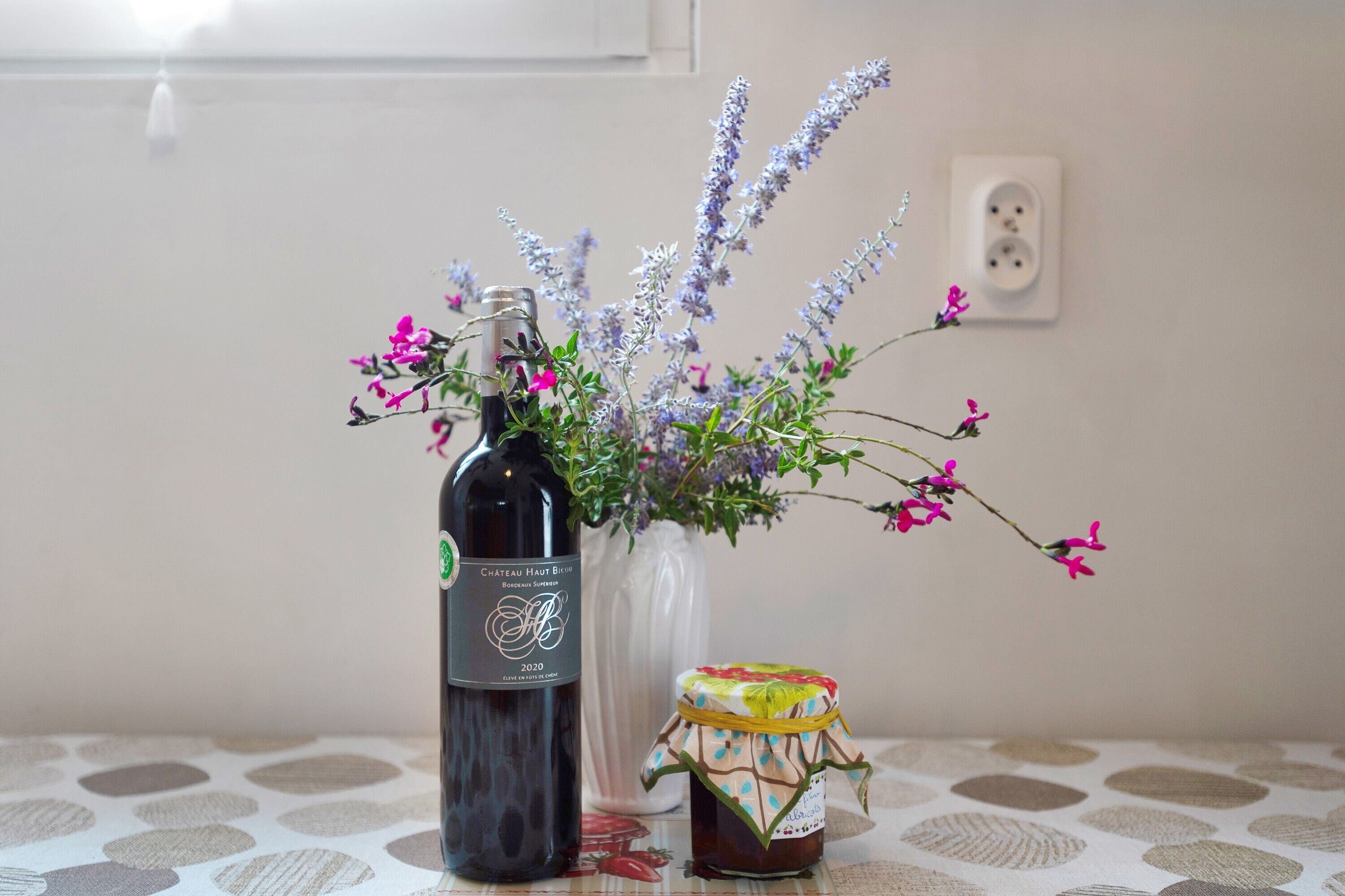 Eine Flasche Chateau Haut Brion 2020 Wein, eine Vase mit lavendelfarbenen und rosa Blumen sowie ein Einmachglas Marmelade auf einem Tisch mit gemustertem Tischläufer.