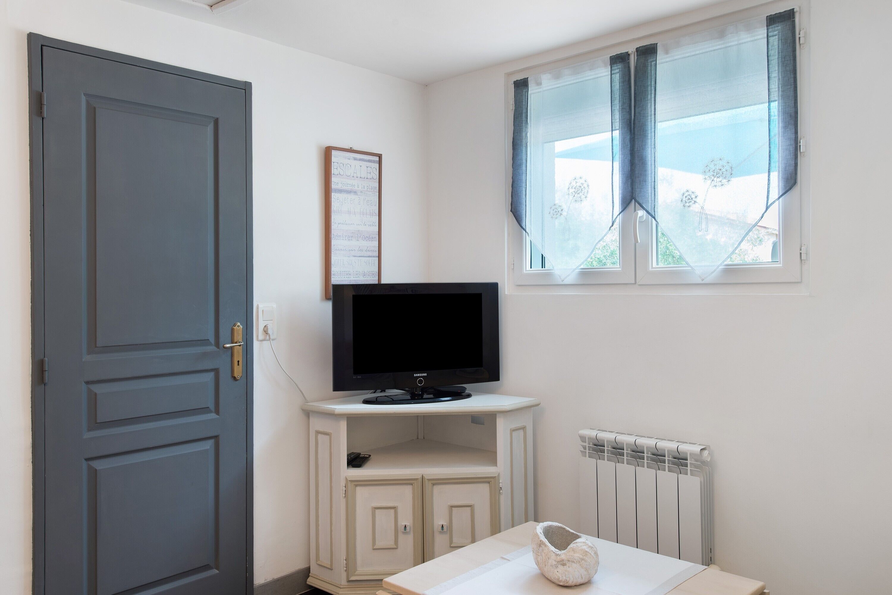 Una habitación con puerta cerrada, televisor montado, radiador y ventana con cortinas.