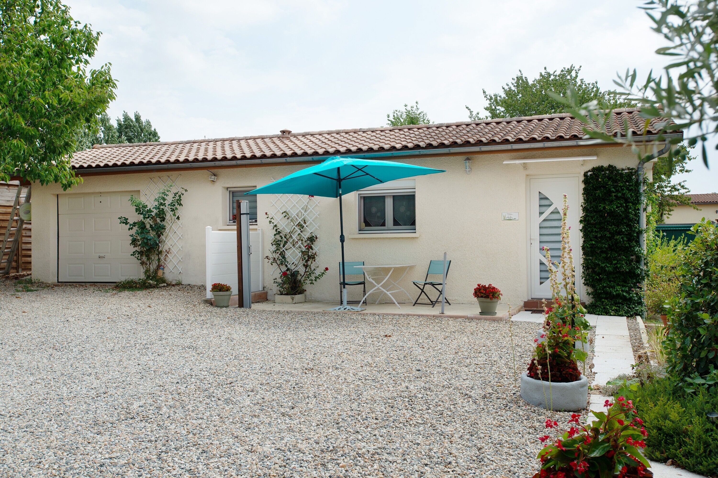 Une maison avec une allée en gravier, une terrasse avec une table et des chaises, et un parasol bleu.