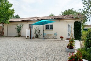 Een huis met een grindpad, een terras met een tafel en stoelen, en een blauwe parasol.
