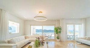 Un salon spacieux avec un grand canapĂ©, une table basse agrĂ©mentĂ©e de plantes et une vue sur la mer grĂące Ă de grandes fenĂȘtres.