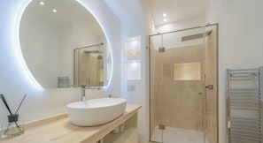 Une salle de bain moderne avec un grand miroir rond, un lavabo blanc, une douche en verre et un porte-serviettes.