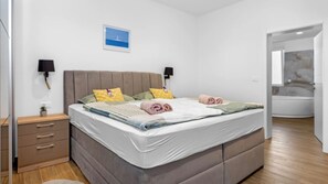 A bedroom with a bed, bedside tables, a nightstand, a wall-mounted lamp, a framed picture, and a bathroom with a bathtub. - Villa Pinus by Villas Guide (Crvar)