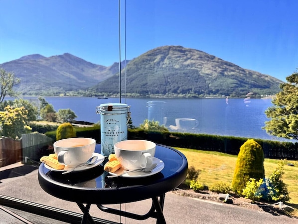Ein Tisch mit zwei Kaffeetassen und Gebäck, ein Einmachglas mit Schokoladenkeksen und Blick auf einen See mit Bergen im Hintergrund.