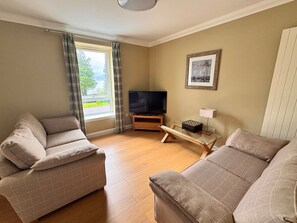 A living room with two sofas, a TV, a glass coffee table, and a window with curtains. - Apartment 1 Lochside with Balcony (Onich by Fort William)