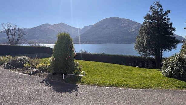A view of a lake with mountains in the background, a well-maintained lawn, and a hedge. - Allt Nan Ros Cottage (By Fort William)