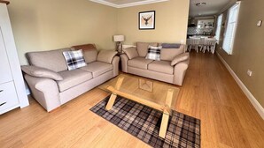 A living room with two beige sofas, a glass coffee table, a plaid rug, and a kitchen in the background. - Allt Nan Ros Cottage (By Fort William)