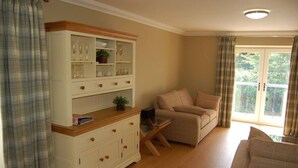 Ein Zimmer mit einer Couch, einem Schrank mit Gläsern, einem Laptop auf einem kleinen Tisch und Blick ins Grüne durch ein Fenster.