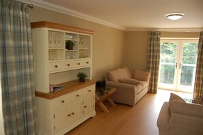 A room with a sofa, a cabinet with glassware, a laptop on a small table, and a view of greenery through a window. - Stables Cottage near Fort William (Onich)