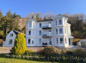 A multi-story building with balconies, surrounded by a garden with daffodils. - Apartment 2 Lochside with Balcony (Onich)